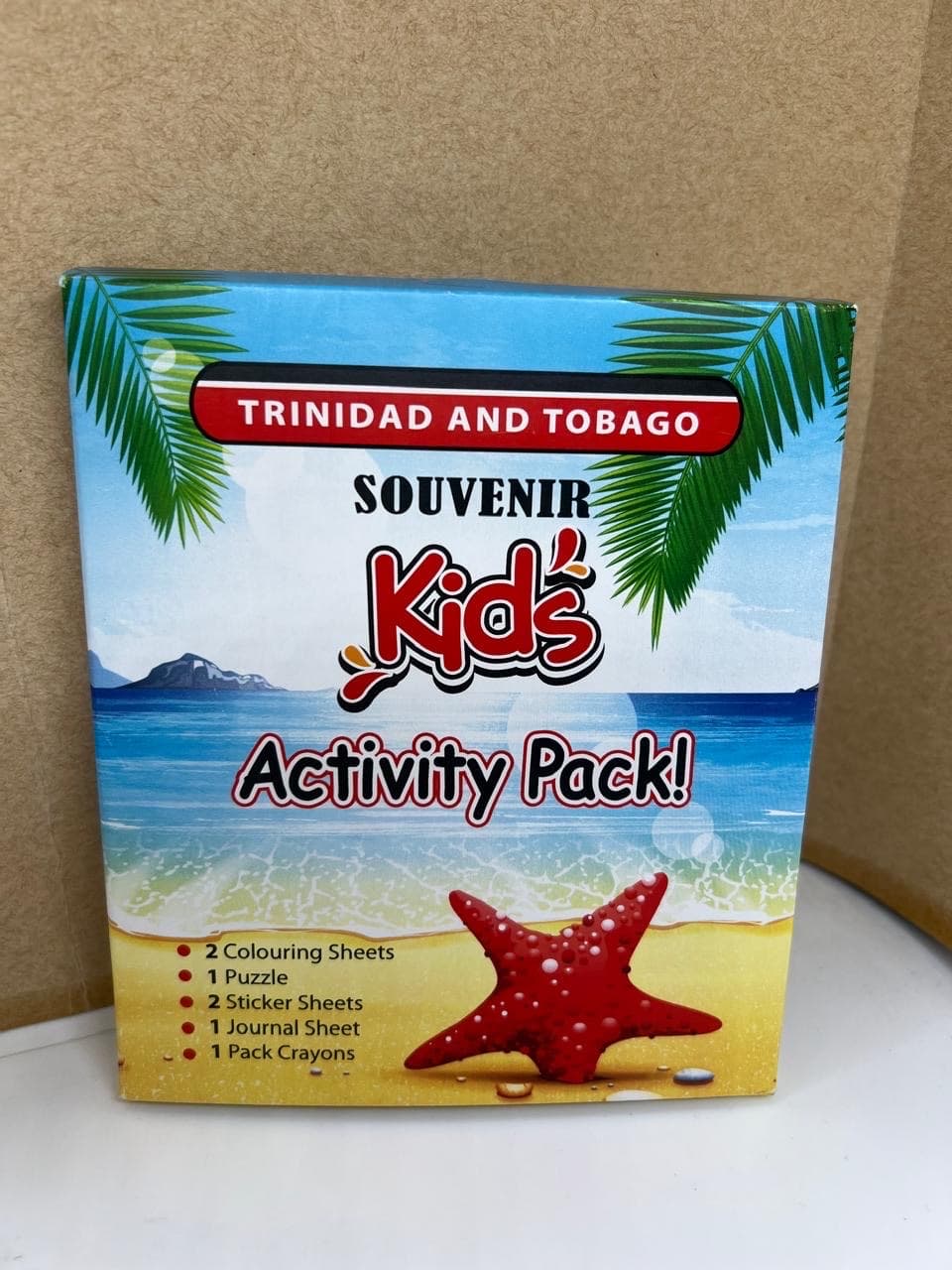 Front of the Trini Souvenir Kids Activity Pack box with a beach, ocean, palm leaves, and red starfish